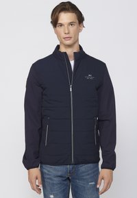 Mørkeblå polstret jakke med høj krave, lynlåslukning foran, kontrastærmer, to lynlåsede lommer og et lille logo på brystet.