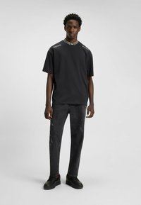Sort kortærmet skjorte med høj krave og grå striber, med "HUGO" branding på skuldrene. Båret med mørke jeans og sorte sko.