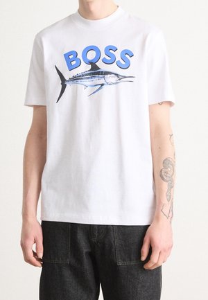 Person iført en hvid t-shirt med blåt marlin-grafik og teksten "BOSS" øverst, kombineret med sorte jeans og synlige armtatoveringer.