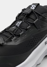 Chaussure de sport noire avec un dessus en maille texturée, un système de laçage élastique et une semelle intermédiaire blanche. Présente un logo sur la languette et sur le côté.