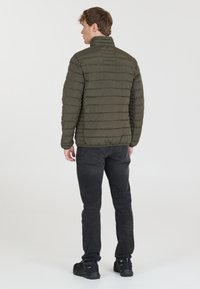 Veste en duvet vert olive avec des coutures horizontales, un col haut et une texture lisse, portée avec un jean foncé et des chaussures noires.