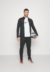 Sort træningsjakke med lynlås, hvid Nike-logo t-shirt, sorte bukser og brune træningssko, holder en grå fodbold.