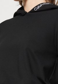 Primer plano de una persona que lleva una sudadera con capucha negra con el logo de Calvin Klein visible en el forro interior de la capucha.