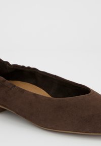 Bruine suède platte schoen met een afgeronde neus en licht geplooide kraag, zichtbaar op een witte achtergrond.