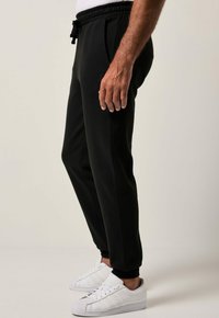 Man in zwarte joggingbroek met een trekkoord in de taille en witte sneakers, staande tegen een effen lichte achtergrond.