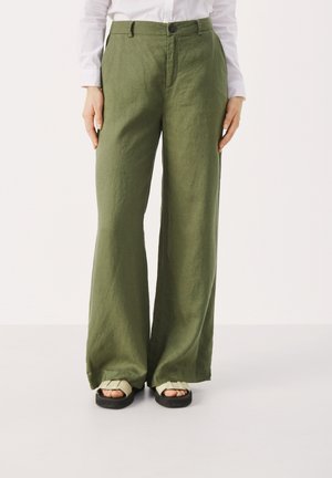 Personne portant un pantalon large vert olive, une chemise blanche à manches longues et des sandales crème avec des semelles noires, debout sur un fond uni.