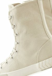 Zapatillas altas de lona beige con ojales de metal y cordones planos, que presentan una superficie texturizada y una suela de goma con un patrón en zigzag.