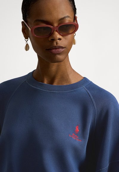 Blå genser med ribbet halslinning og raglanermer, med en liten rød brodert logo på brystet, brukt med rosa solbriller.