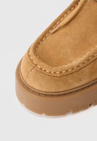 Tan suede kengät paksulla kumipohjalla, näkyvillä ompeleilla ja nauhakiinnityksellä, valokuvattu valkoista taustaa vasten.