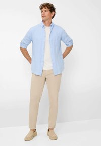 Camicia azzurro chiaro con bottoni sopra una t-shirt bianca, abbinata a pantaloni beige e scarpe slip-on marrone chiaro. Insieme semplice e casual.