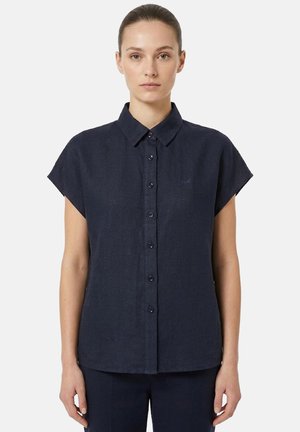 Donna con capelli raccolti, indossa una camicia blu navy a maniche corte con bottoni e pantaloni abbinati dello stesso colore, in piedi davanti a uno sfondo uniforme.