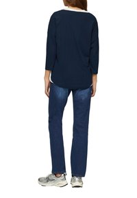 Marineblauw ribbelshirt met driekwartmouwen, losse pasvorm, gedragen over een wit hemd. Gecombineerd met donkerblauwe jeans en zilveren sneakers.