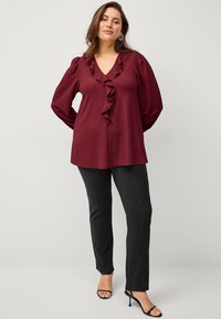 Bordeauxfarbene Bluse mit rüschigem V-Ausschnitt, strukturierter Stoff und langen Ärmeln, kombiniert mit schwarzen, figurbetonten Hosen und Riemchen-Heels.