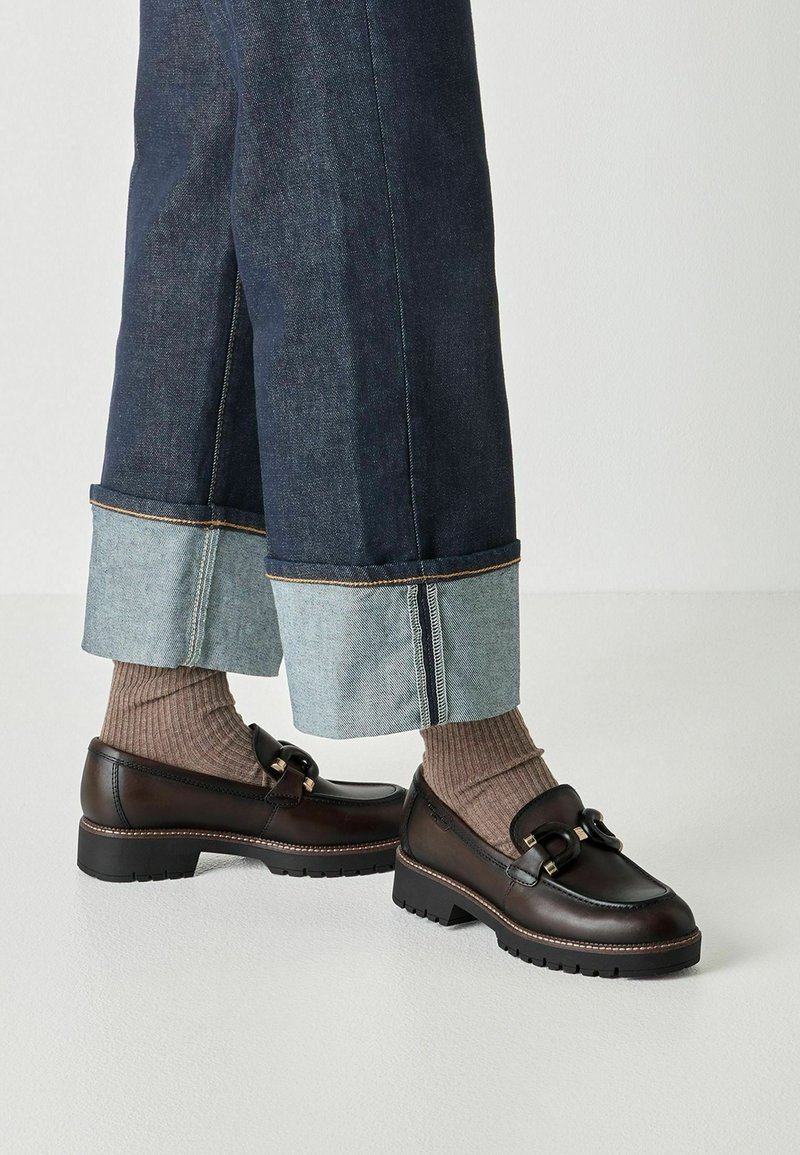 Σκούρα καφέ δερμάτινα loafers με χοντρές σόλες, φορεμένα με μπεζ ριμπ καλτσάκια και φαρδιά, γυριστά σκουρόχρωμα τζιν, σε λευκό φόντο.