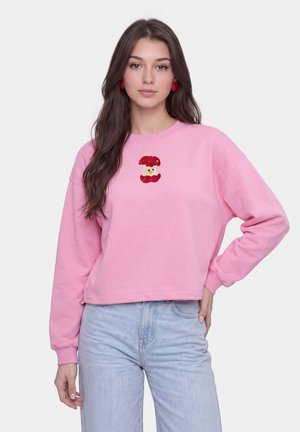 Vrouw met lang bruin haar die een roze trui draagt met een klein rood appelklokhuisontwerp en lichtblauwe spijkerbroek, staand met één hand op haar heup.