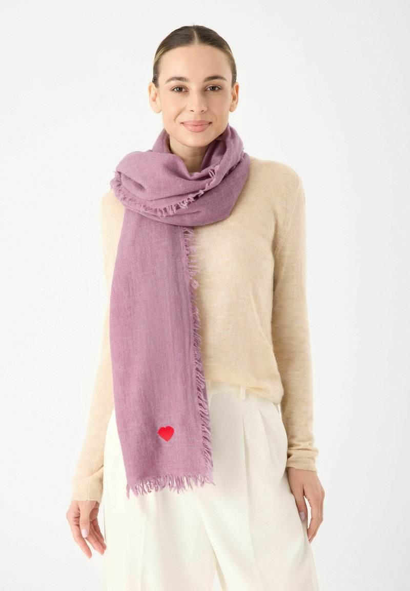 Écharpe violette avec des bords effilochés, arborant un petit détail en forme de cœur rouge. Portée par-dessus un pull beige clair, créant un contraste de textures douces.