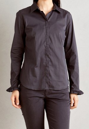 Femme portant une chemise ajustée gris foncé à manches longues boutonnée avec des poignets à volants et un pantalon gris foncé assorti sur un fond uni.