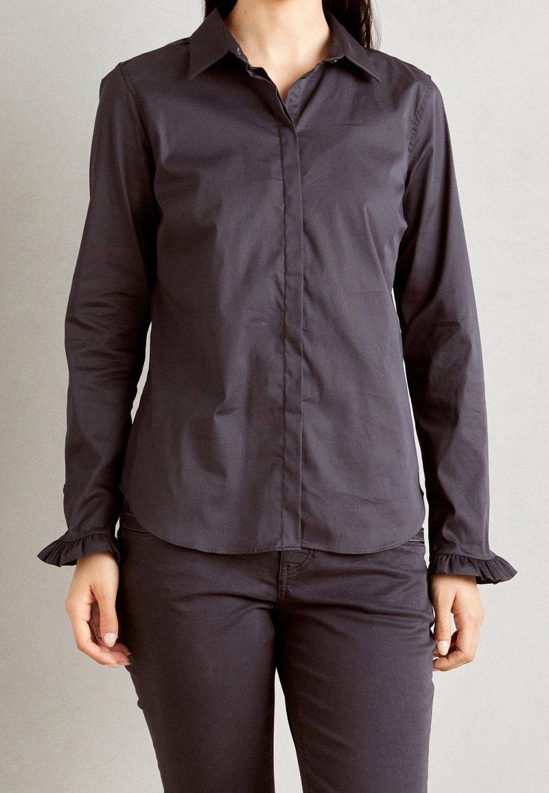 Femme portant une chemise ajustée gris foncé à manches longues boutonnée avec des poignets à volants et un pantalon gris foncé assorti sur un fond uni.