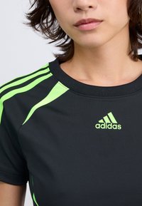 Schwarzes Sportshirt mit grünen Akzenten, ausgestattet mit drei grünen Streifen auf den Schultern und einem grünen Adidas-Logo auf der Brust.
