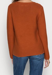Rust-colored knitted sweater with a textured finish, featuring long sleeves and a rounded neckline, paired with blue denim bottoms.