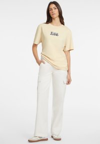 Jasnożółta koszulka z grafiką "JEANS", w połączeniu z białymi spodniami cargo z bocznymi kieszeniami, noszona z beżowo-białymi sneakersami.