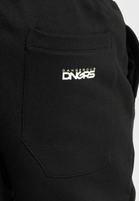 Schwarze Baumwoll-Sweatpants mit einer Gesäßtasche, auf der der Text "DANGEROUS DNGRS" in Weiß und neongelb zu sehen ist. Der Text ist fett und zentriert.