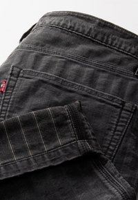 Pantalones vaqueros negros con textura de espiga, costuras en contraste y bolsillos visibles. Incluyen una etiqueta de la marca en la cintura.