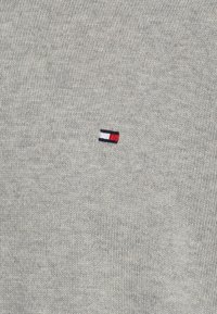 Grau gestrickter Pullover mit einem kleinen, in Rot, Weiß und Blau gestickten Logo auf der Brust. Das Material hat eine weiche, strukturierte Oberfläche.