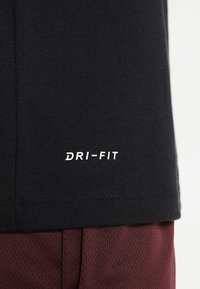 Svart Dri-Fit T-shirt har en jämn textur, med en vit "DRI-FIT" logotyp på den nedre fållen. Positionerad ovanför vinröda shorts.
