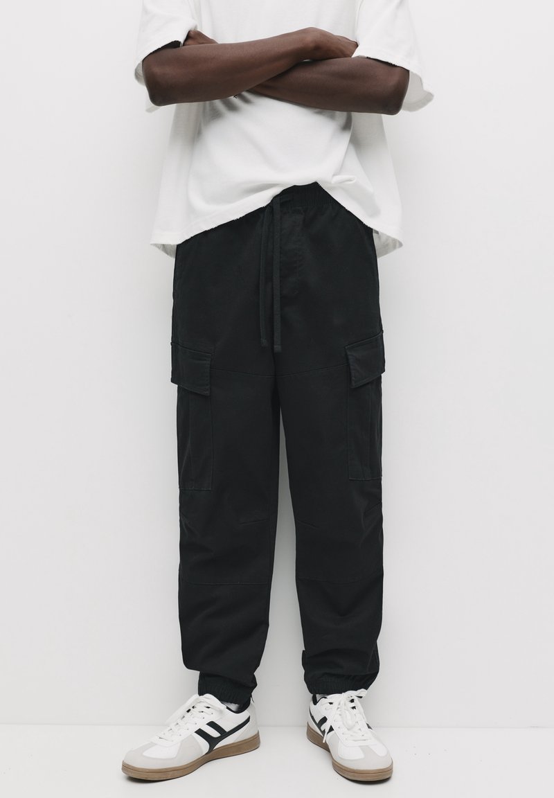 Personne les bras croisés portant un pantalon cargo noir, des baskets blanches avec des bandes noires et une chemise blanche ample à manches courtes.