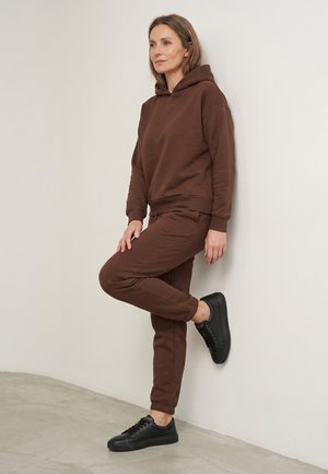 Frau lehnt an der Wand, trägt braunen Hoodie, passende Jogginghose und schwarze Sneakers, schaut leicht zur Seite mit neutralem Gesichtsausdruck.