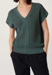 Donna che indossa un top a maglia a maniche corte color teal con scollatura a V e pantaloni neri, mani nelle tasche, su uno sfondo chiaro uniforme.