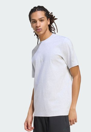 Jonge persoon met dreadlocks, gekleed in een lichtgrijs T-shirt met korte mouwen en een zwarte broek, staand tegen een effen lichte achtergrond.