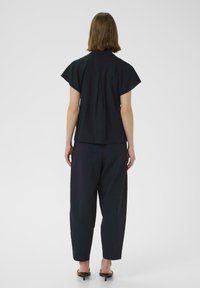 Blusa nera a maniche corte con retro plissettato, abbinata a pantaloni larghi scuri. Il tessuto sembra leggero e liscio.