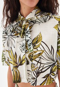 Blusa bianca a maniche corte con colletto e farfallino, caratterizzata da motivi di foglie verdi e marroni. Dettagli con bottoni sul pannello anteriore.