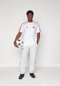 Biela športová dres so červenými pruhmi a logom, skombinovaný so svetlošedými teplákmi. Čierny futbalový loptú drží v jednej ruke, čierne tenisky.