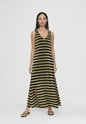 Abito lungo con strisce verticali nere e oro, scollatura a V, design senza maniche e una silhouette fluida. Indossato con sandali neri a infradito.