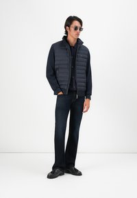 Mørk puffervest med glidelås over en marineblå skjorte, kombinert med tettsittende mørke jeans og svarte sko. Tilbehørt med solbriller.