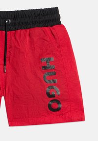 Κόκκινα μαγιό σορτς με μαύρη ελαστική μέση και κορδόνι, με μεγάλο κάθετο κείμενο "HUGO" στο αριστερό πόδι.