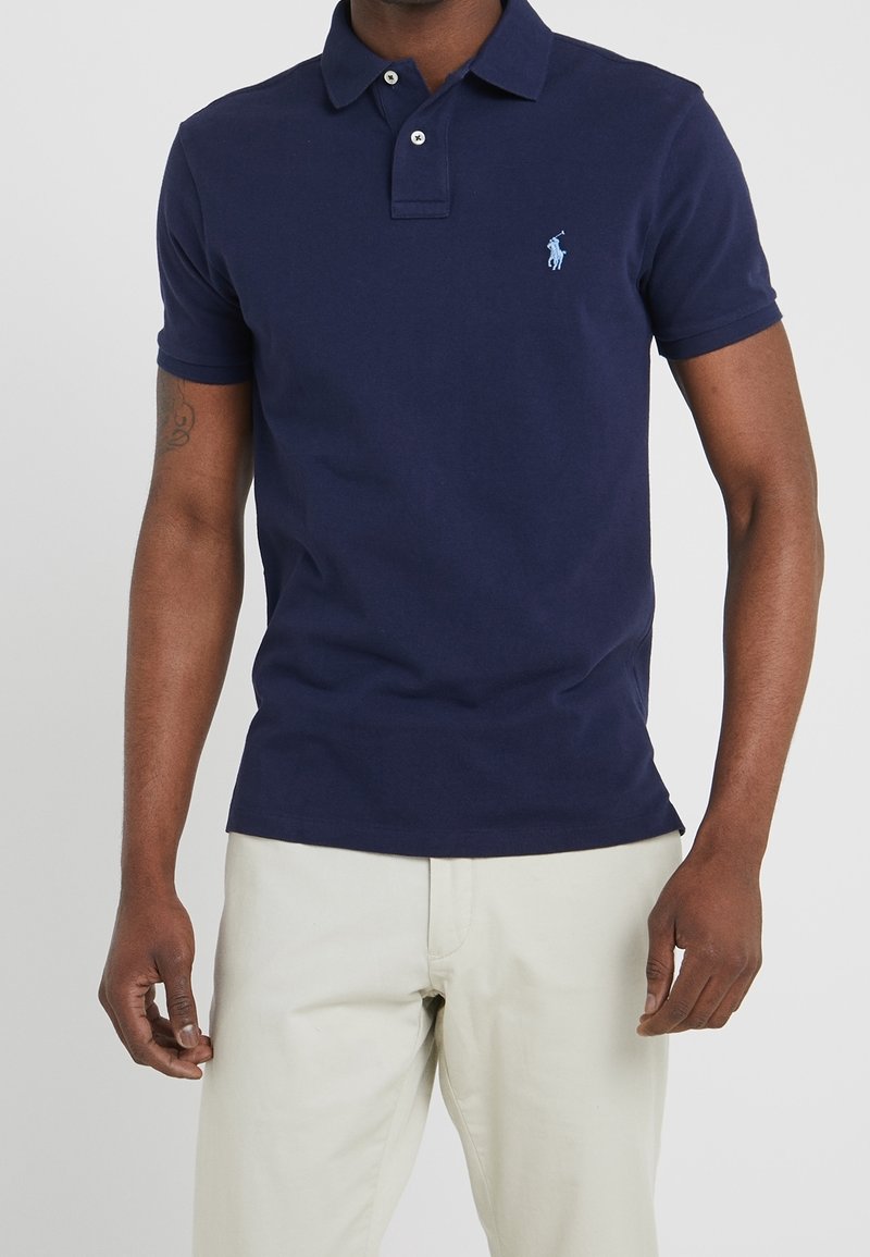 Mann in einem taillierten navyblauen Poloshirt mit kleinem hellblauen Logo auf der Brust und offweißen Hosen vor einem einfarbigen Hintergrund.