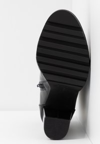 Semelle d'une botte en cuir noir avec des rainures horizontales et un talon bloc, positionnée à la verticale contre un mur d'angle blanc.