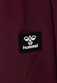 Burgunderfarbener Stoff mit einem schwarzen rechteckigen Patch, auf dem ein weißes Logo und das Wort "hummel" am unteren Rand geprägt sind. Glatte Textur.