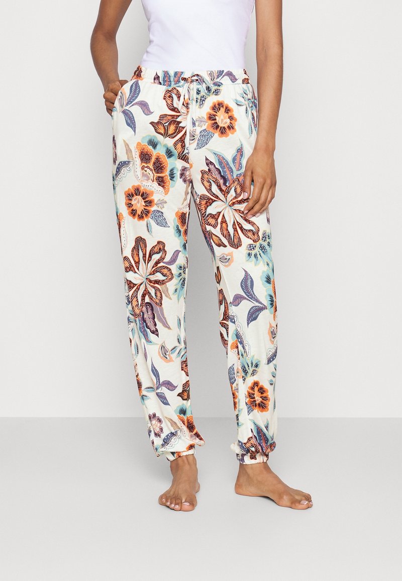 Femme portant un pantalon blanc à cordon avec de grands motifs floraux orange, bleu et violet, associé à un top blanc sans manches.