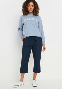 Lichtblauwe sweatshirt met de tekst "WELLNESS", gecombineerd met donkerblauwe cropbroeken. De outfit wordt aangevuld met witte sneakers.