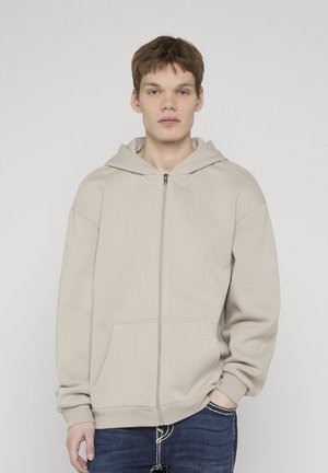 Young man with short brown hair wearing a beige zip-up hoodie and dark blue jeans, standing against a plain light background.