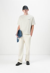 Locker sitzendes, hellgraues T-Shirt, beigefarbene Hose und schwarze Schuhe; hält eine blaue Jeansjacke. Minimalistisches Design mit subtiler Markenkennzeichnung.