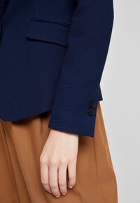 Blazer texturizado azul marino con un corte ajustado, que presenta un bolsillo y dos botones negros en la muñeca, combinado con pantalones de tela marrones.