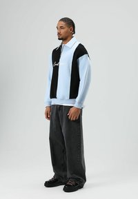 Sweat-shirt bicolore bleu clair et noir avec une fermeture éclair, arborant un texte brodé. Associé à un jean ample foncé et des chaussures noires épaisses.