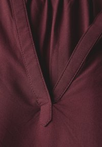 Tessuto bordeaux con design a profondo scollo a V. Presenta pieghe e un accento a punta all'linea del collo, che mette in evidenza una superficie liscia e cuciture pulite.