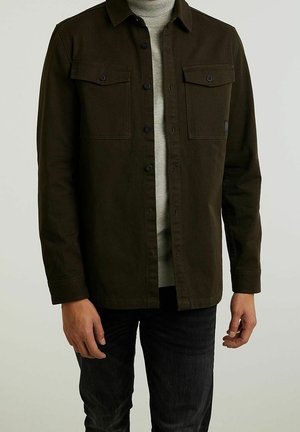 Man wearing dark brown button-up jacket over light gray turtleneck sweater and black pants, standing against plain background.